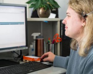 eine blonde Frau mit Headset guckt auf einen Computerbildschirm